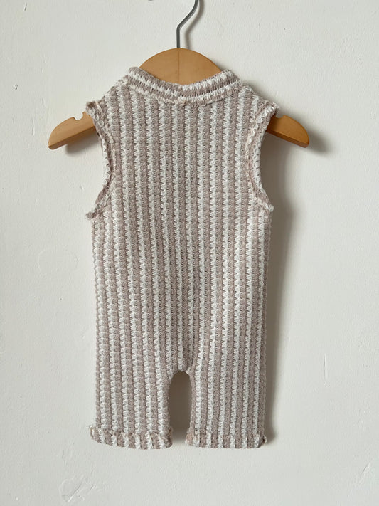 back of a sleeveless knit romper in a textured waffle pattern of beige and white, hanging on a wooden hanger against a plain off-white wall.