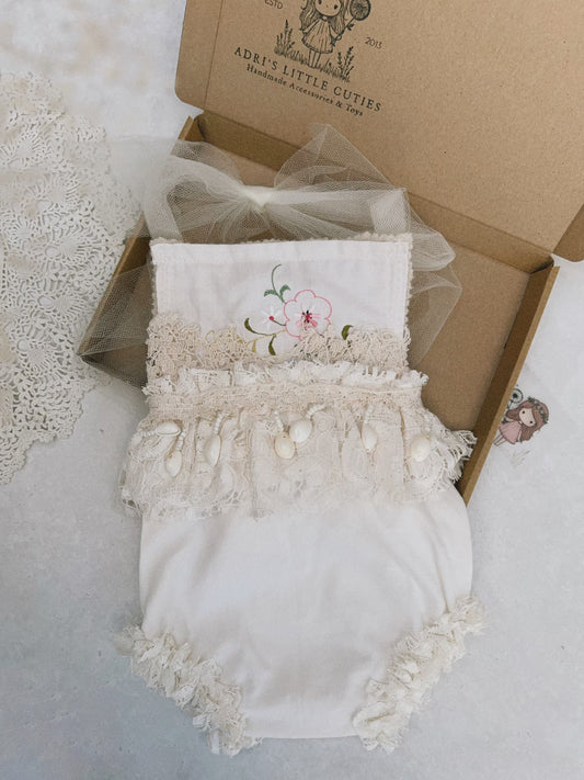 A cream-colored baby romper with floral embroidery, lace trim, and pearl embellishments, presented in a brown cardboard gift box with a bow. The box is branded 'ADRI'S LITTLE CUTIES'.
