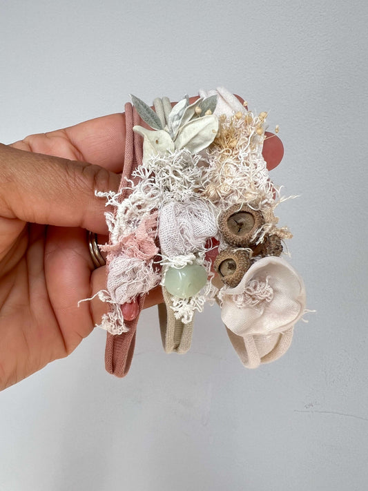 A hand holds three delicate fabric headbands decorated with lace, dried flowers, buttons, and a small green bead, against a plain light gray wall.