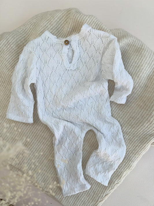 A white long-sleeve baby romper with a textured knit pattern and a small wooden button closure at the collar, laid flat on a beige knit blanket.