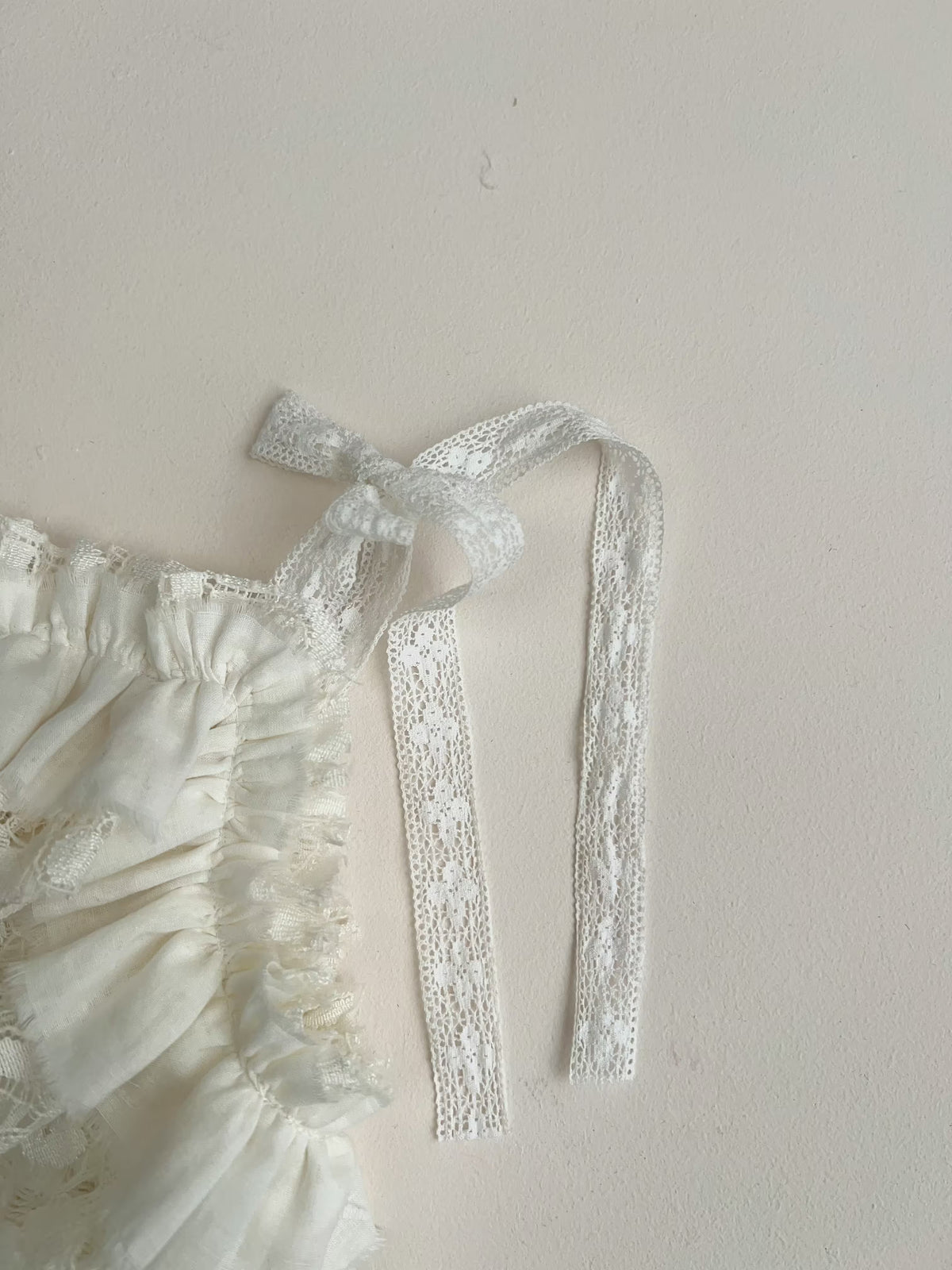 White lace ribbon tie with a bow and long trailing ribbon, attached to a ruffled cream fabric garment, shown against a plain off-white wall.