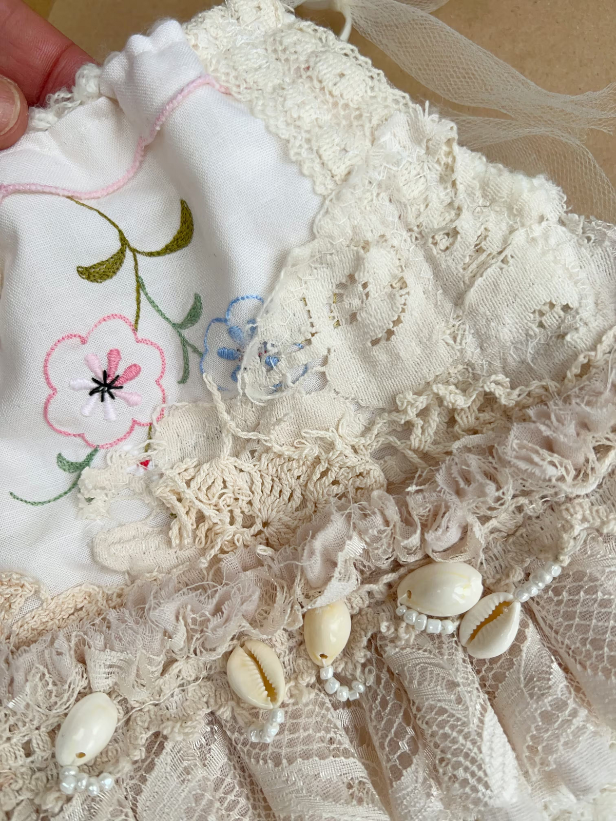 A close-up of a handmade bohemian-style baby garment or textile piece featuring cream lace, cowrie shells, tulle, and white fabric with pink and green floral embroidery.