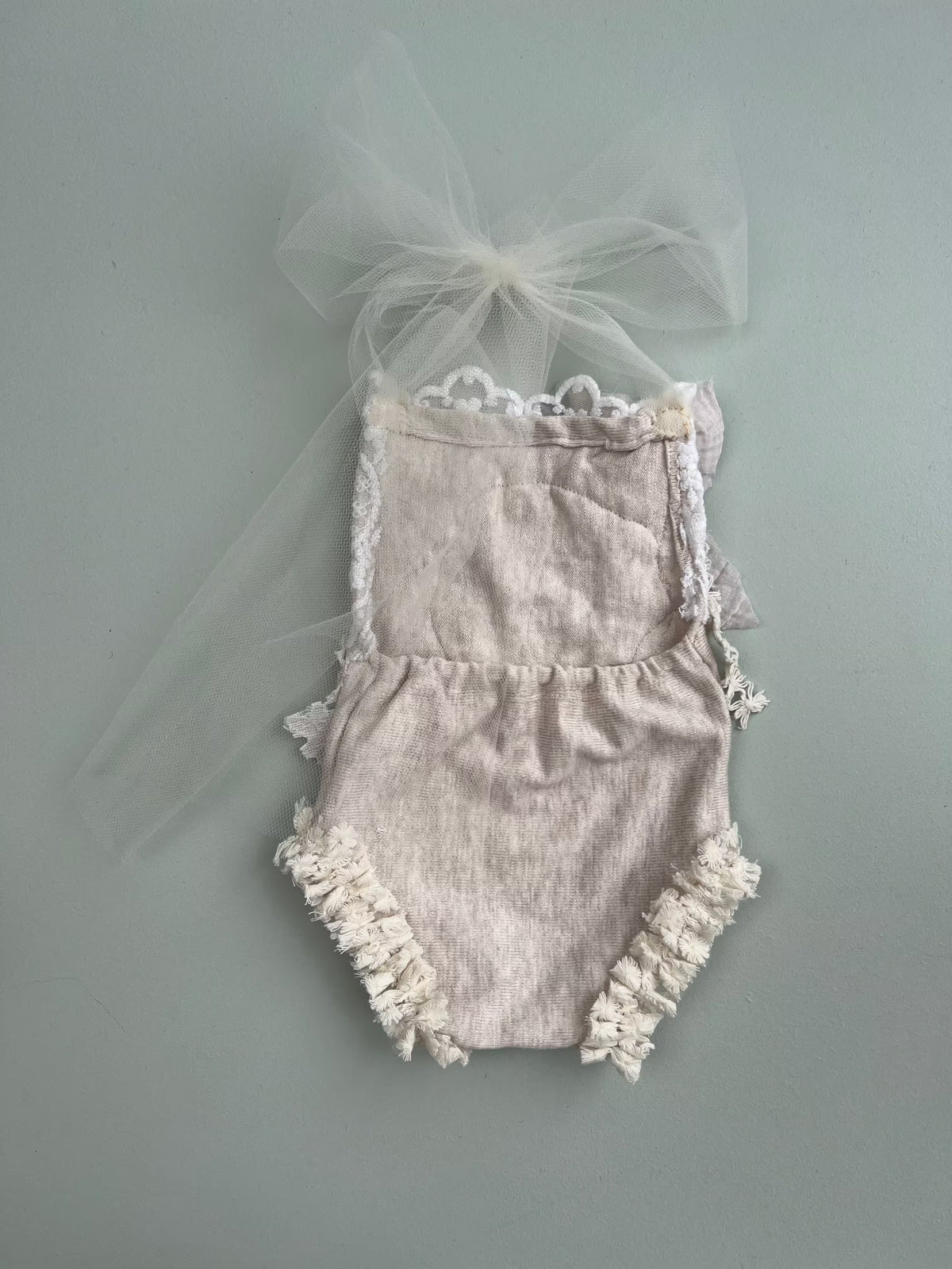 detail of the back of the rompers,  baby romper in a light beige linen-like fabric with white lace trim, featuring a ruffled hem and a matching sheer white tulle bow on the back, laid flat on a muted gray-green background.
