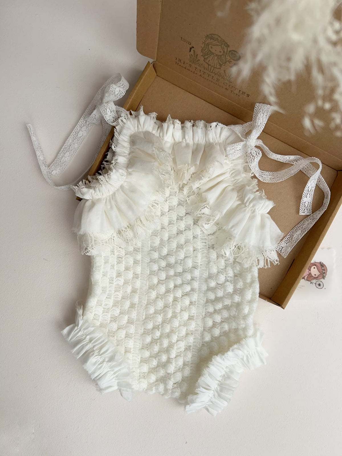 A white textured romper with ruffled trim and lace tie straps, laid flat on a light beige surface next to an open brown gift box.