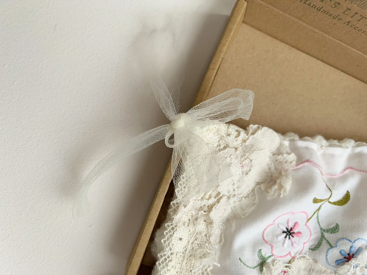 A close-up of a white embroidered fabric accessory with lace trim and a sheer white ribbon bow, placed beside a brown kraft paper box on a light neutral background.