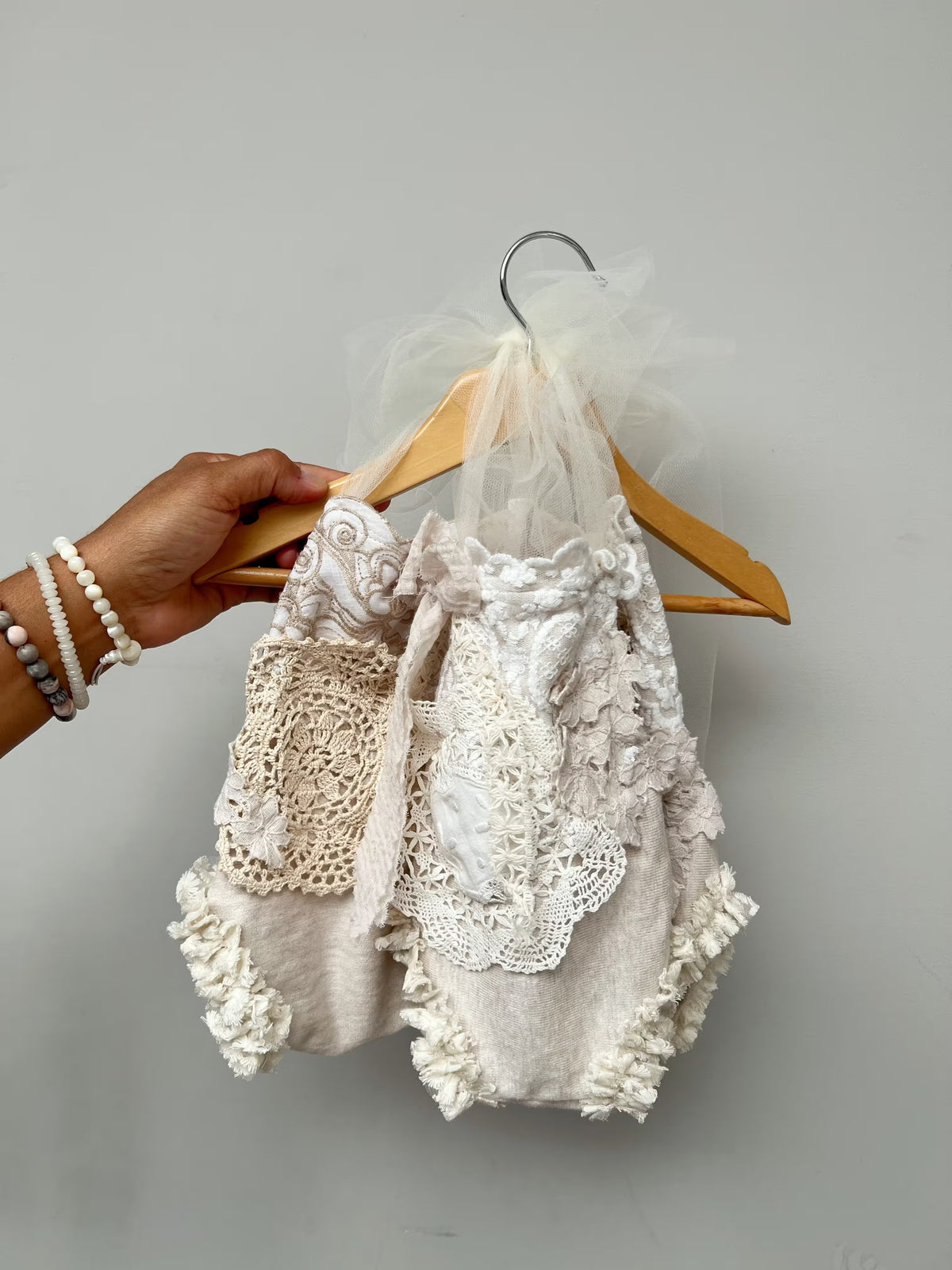 A baby girl's ruffled romper in cream and white lace, held on a wooden hanger by a person's hand against a plain gray background.