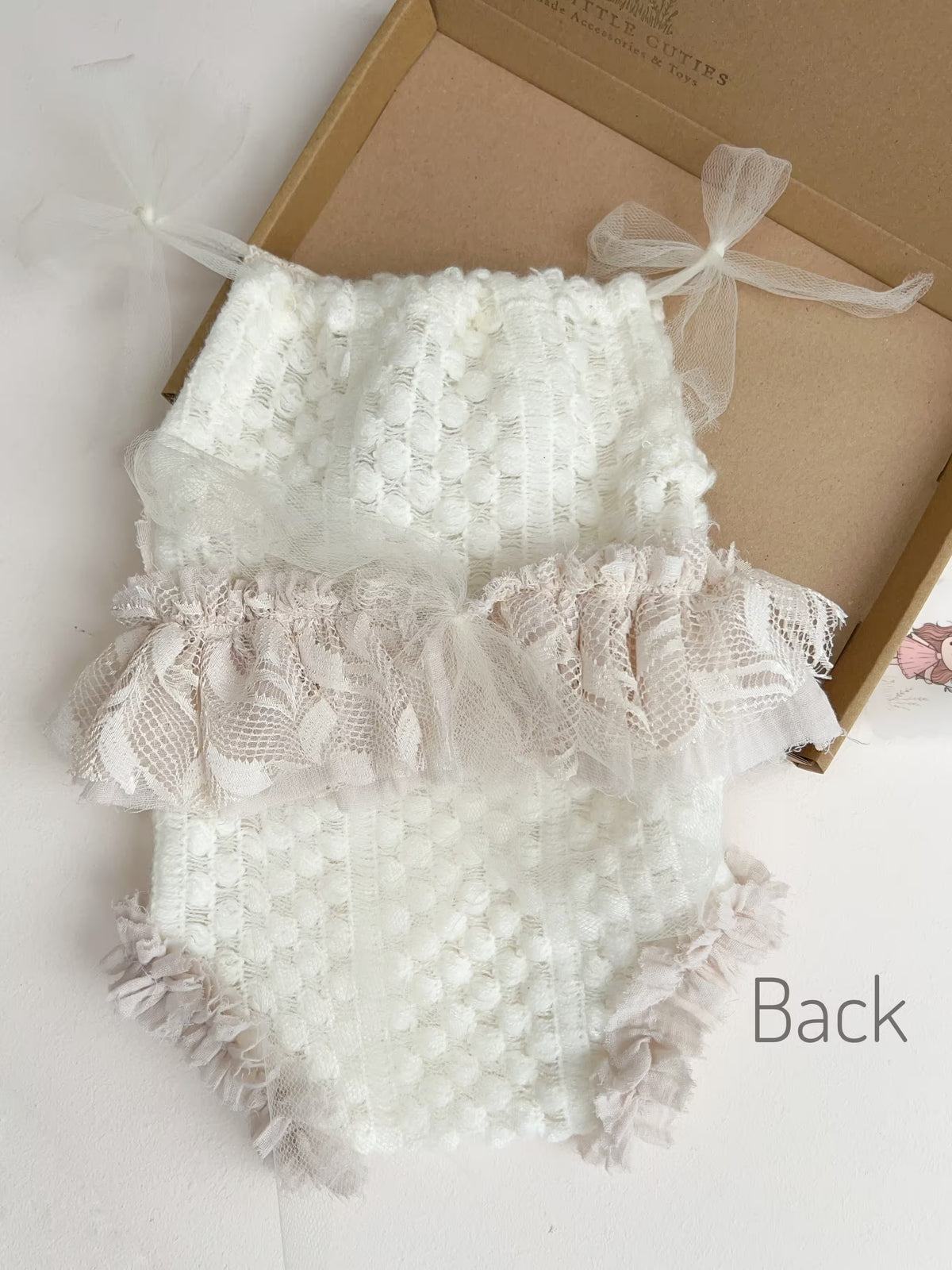 White textured romper with lace ruffles and sheer ribbon bows, displayed on a light surface next to a brown box with the word 'Back' printed on the bottom right.