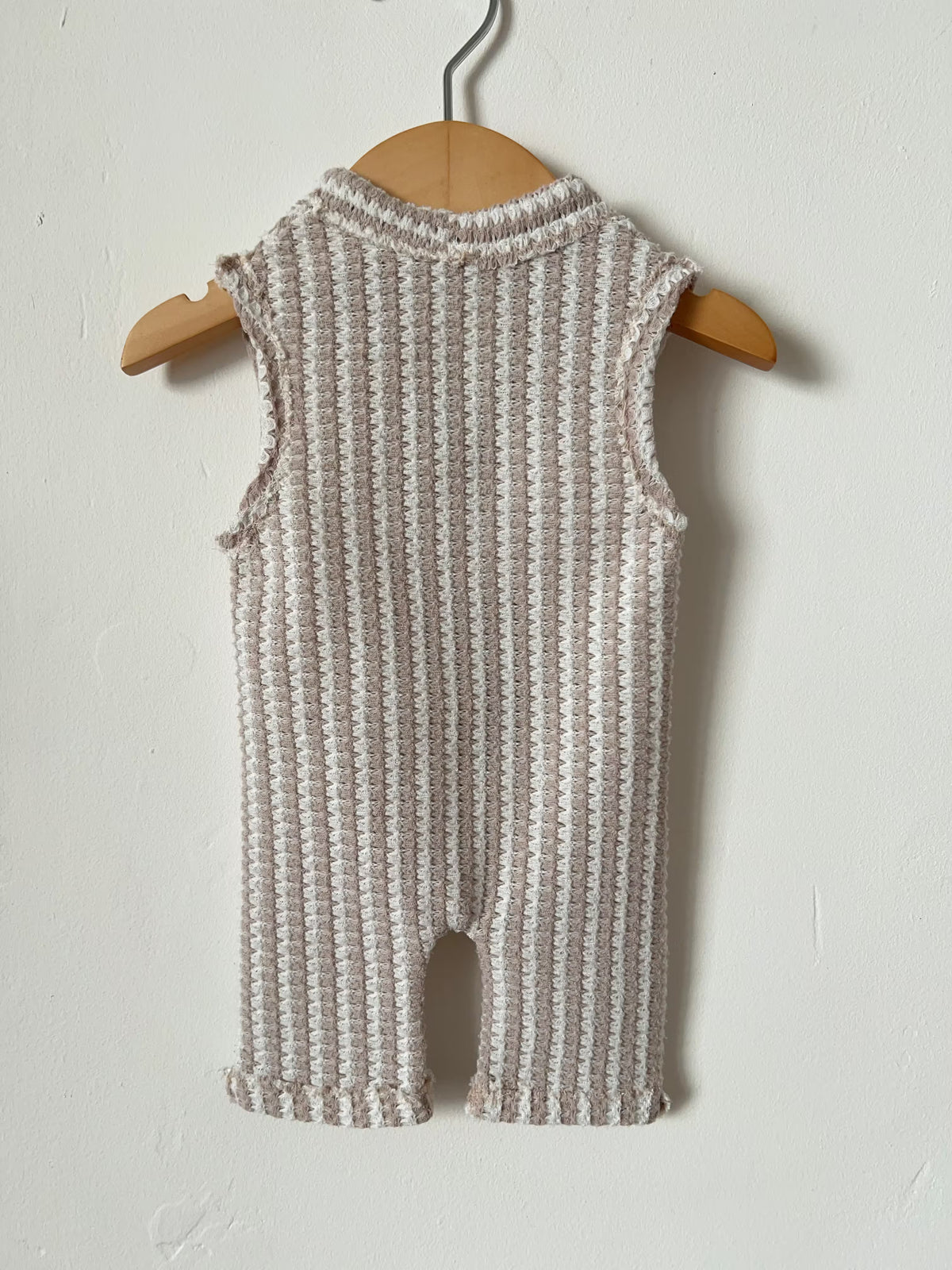back of a sleeveless knit romper in a textured waffle pattern of beige and white, hanging on a wooden hanger against a plain off-white wall.