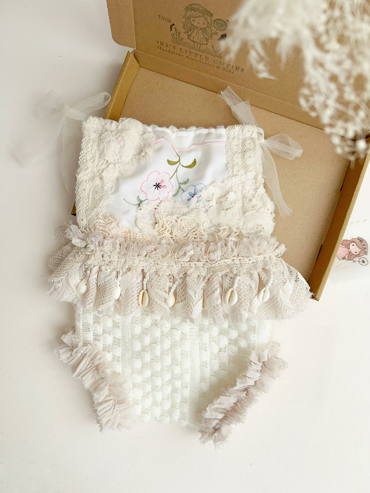 A white and beige baby romper with lace, tulle, and shell embellishments, displayed in an open kraft gift box on a white surface.