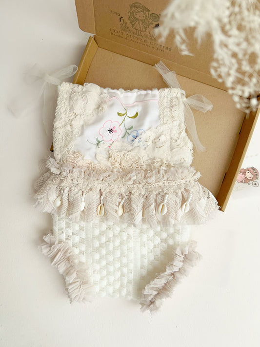 A white and beige baby romper with lace, tulle, and shell embellishments, displayed in an open kraft gift box on a white surface.