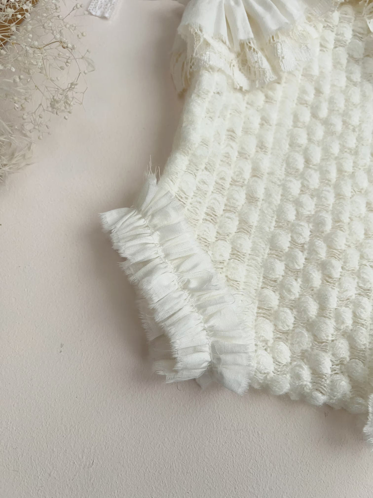 A close-up of a white textured knit garment with ruffled trim, laid flat on a light beige surface.