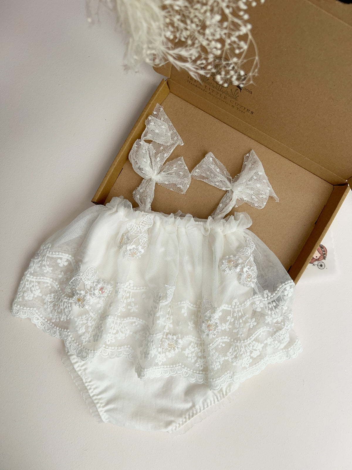 White lace baby bloomers with matching bow headband, displayed inside an open brown cardboard box on a light beige surface.