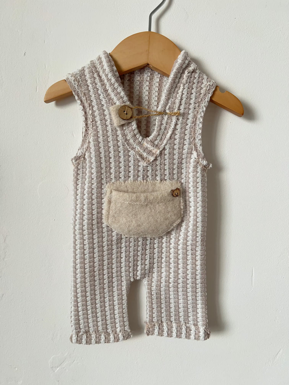 A sleeveless knit baby romper in a textured cream and beige pattern, hanging on a wooden hanger against a white wall. The romper features a soft beige patch pocket on the front with a small wooden button closure.