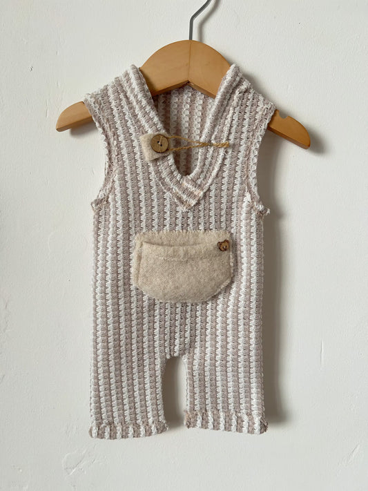A sleeveless knit baby romper in a textured cream and beige pattern, hanging on a wooden hanger against a white wall. The romper features a soft beige patch pocket on the front with a small wooden button closure.