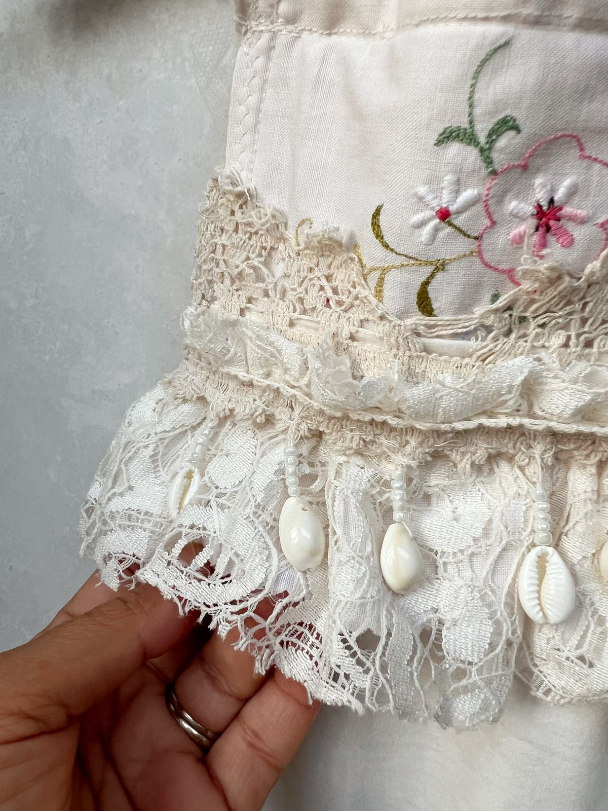 A close-up of a cream-colored embroidered textile with lace trim, shell charms, and floral embroidery, held by a hand.