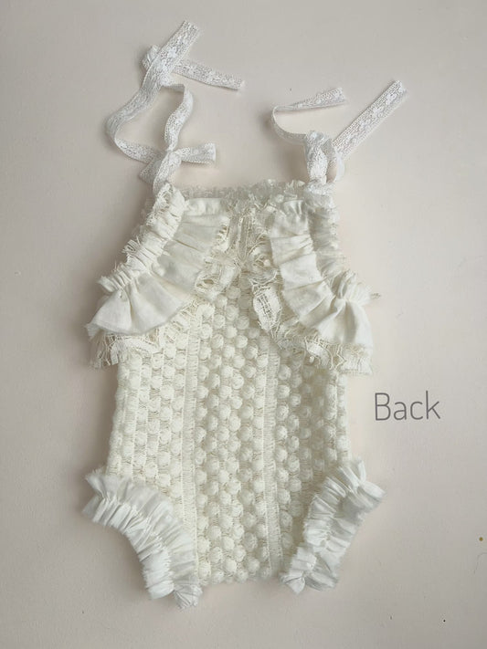 An off-white textured crochet baby bodysuit with ruffle trim and tie straps, shown from the back on a light beige background with the word 'Back' printed on the right.