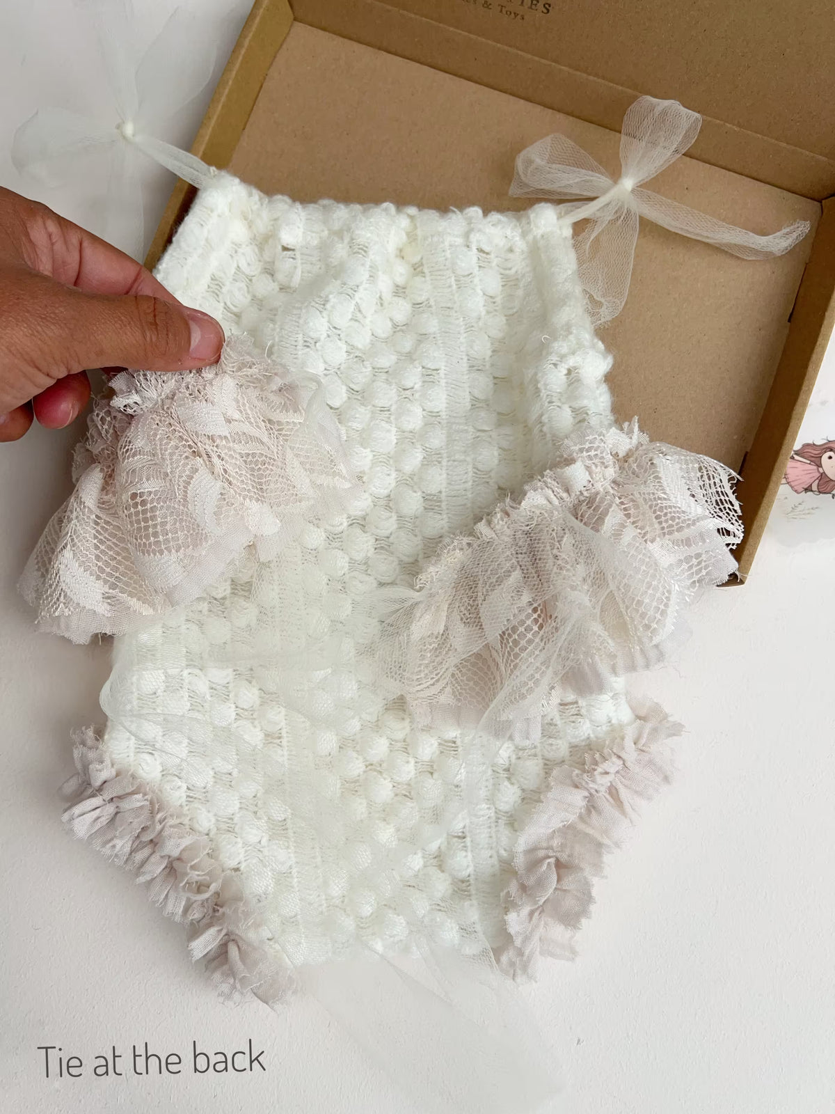 A white textured baby romper outfit and sheer ribbon ties to tie at the back, held by a hand over an open kraft gift box.