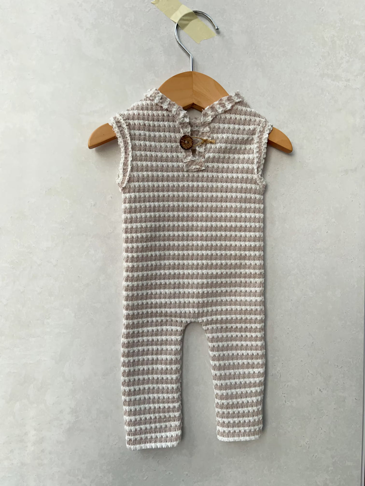 A sleeveless baby romper with a textured waffle-knit pattern in beige and white, featuring a ruffled collar and a wooden button closure, hanging on a wooden hanger against a textured gray wall.