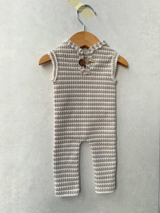 A sleeveless baby romper with a textured waffle-knit pattern in beige and white, featuring a ruffled collar and a wooden button closure, hanging on a wooden hanger against a textured gray wall.