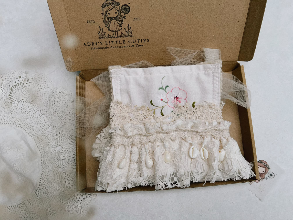 A baby romper in cream lace with a pink floral embroidery presented in a brown kraft paper box on a white background.