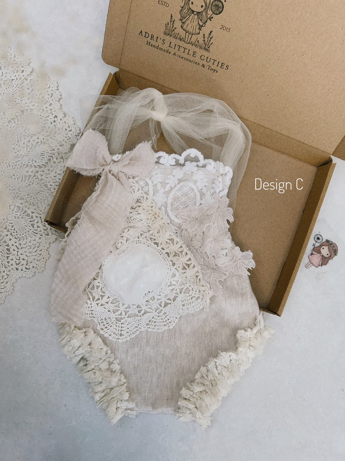 A baby romper in a soft beige linen look  fabric with white lace appliques, a matching bow, and a sheer tulle details, presented in an open brown cardboard box labeled 'ADRI'S LITTLE CUTIES'