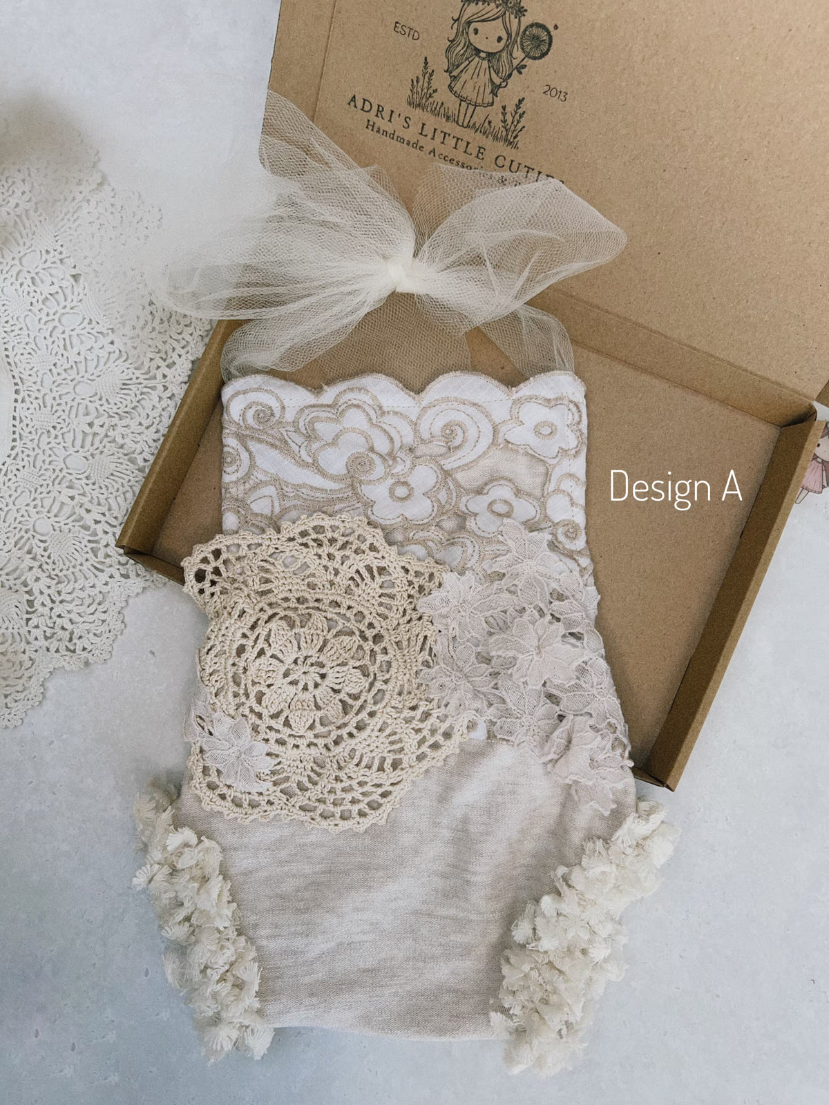 A baby bodysuit in a soft cream or off-white fabric, decorated with intricate floral lace applique, a large crocheted lace patch, and fluffy tassel trim. The bodysuit is styled inside an open kraft paper gift box with a sheer tulle bow, on a light textured background.