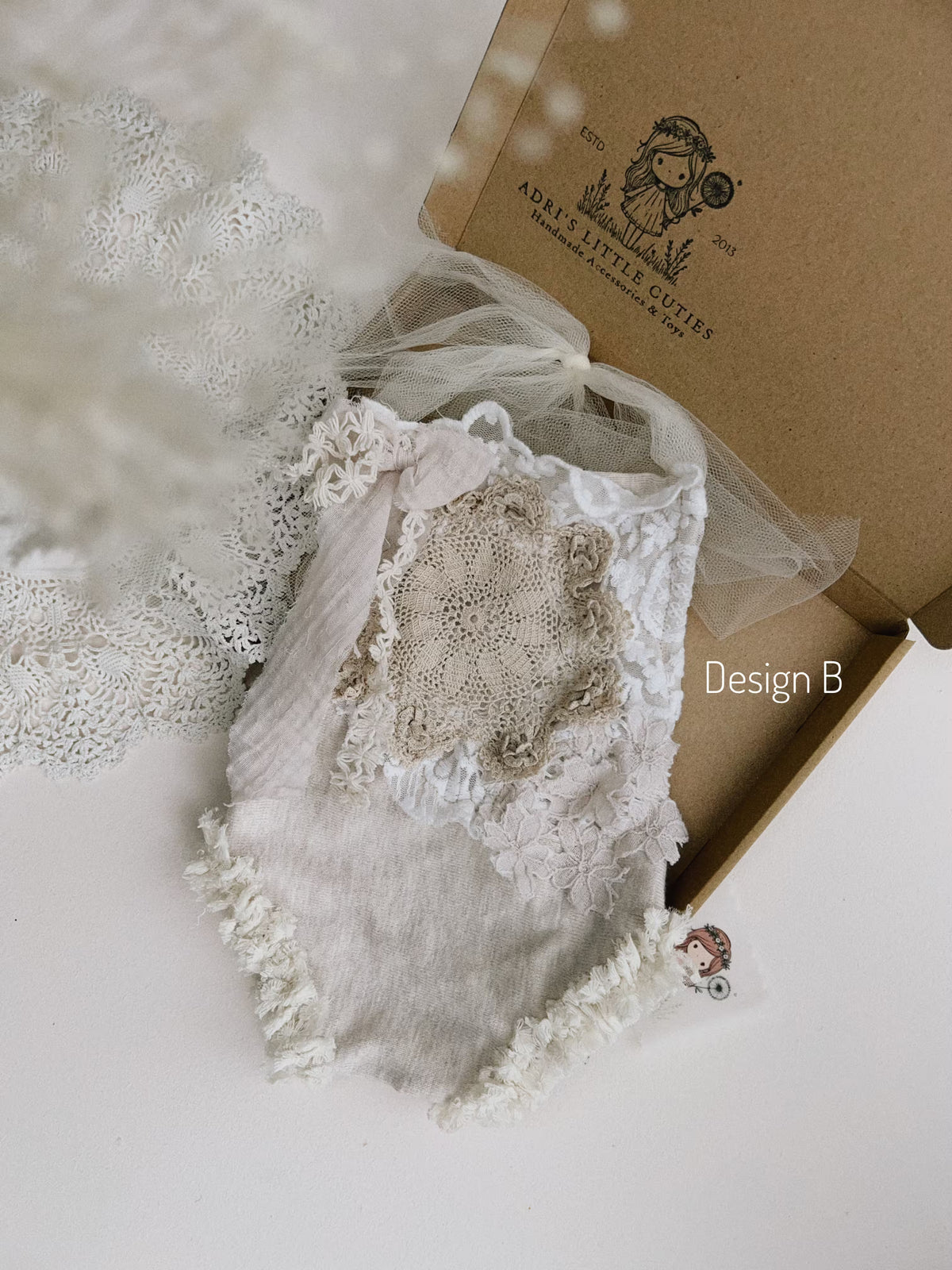 A handmade baby bodysuit in a soft beige and off-white colour palette, featuring textured ruffles, lace applique, a central crochet patch, and a sheer tulle bow on the hanger, set against white window shutters.