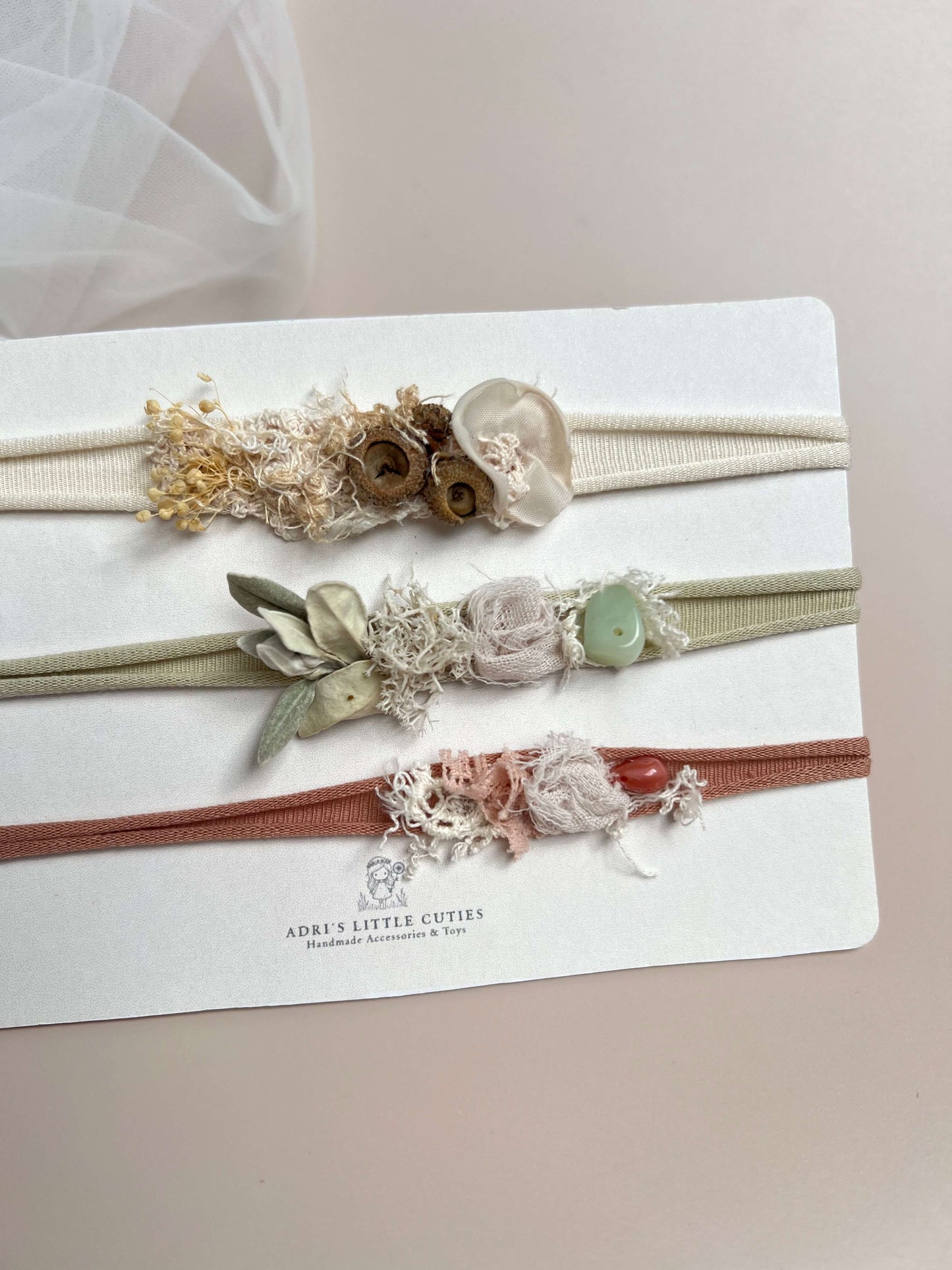 Three delicate fabric headbands displayed on a white branded card. The top headband is cream with dried floral elements and wooden beads; the middle is sage green with faux flowers and a light green stone; the bottom is dusty rose with lace and a red bead.