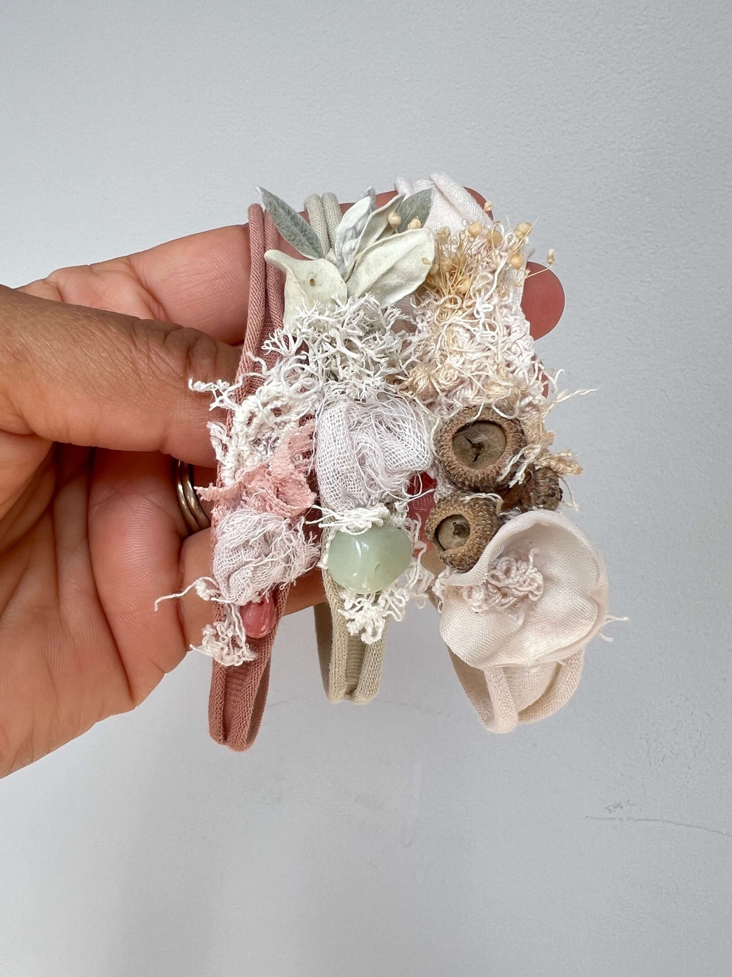 A hand holds three delicate fabric headbands decorated with lace, dried flowers, buttons, and a small green bead, against a plain light gray wall.