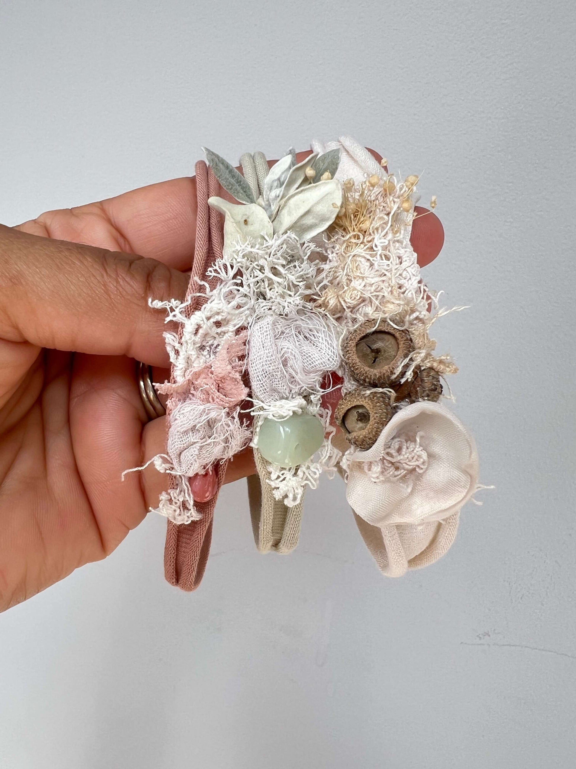 A hand holds three delicate fabric headbands decorated with lace, dried flowers, buttons, and a small green bead, against a plain light gray wall.