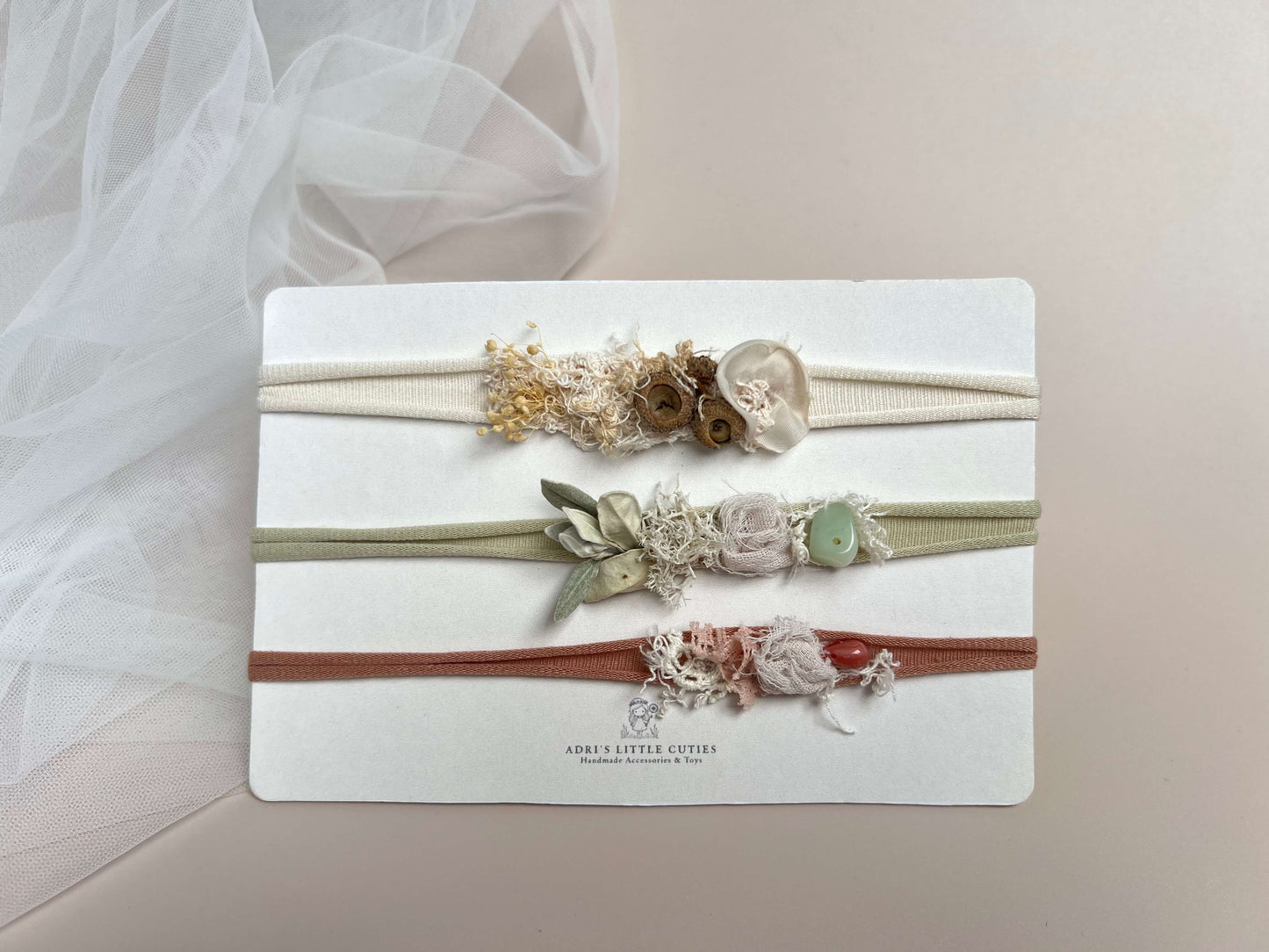 Three delicate floral headbands on a white display card. The top headband is cream with dried beige and brown flowers; the middle is sage green with pale pink and white flowers and a light green bead; the bottom is terracotta red with pink and white flowers.