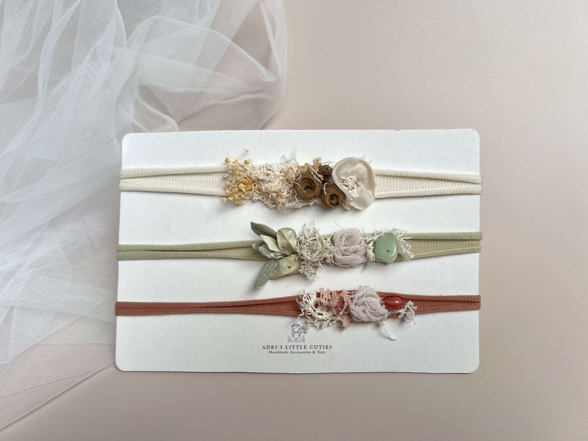 Three delicate floral headbands on a white display card. The top headband is cream with dried beige and brown flowers; the middle is sage green with pale pink and white flowers and a light green bead; the bottom is terracotta red with pink and white flowers.