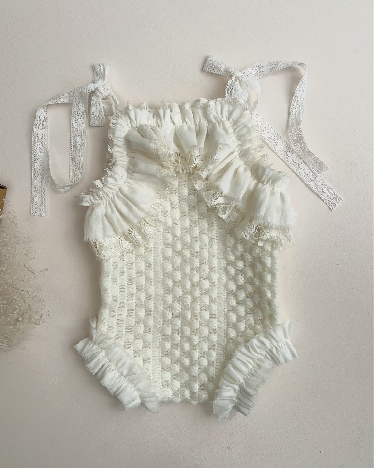 An off-white textured knit baby bodysuit with ruffled trim and shoulder ties, laid flat on a light beige background.
