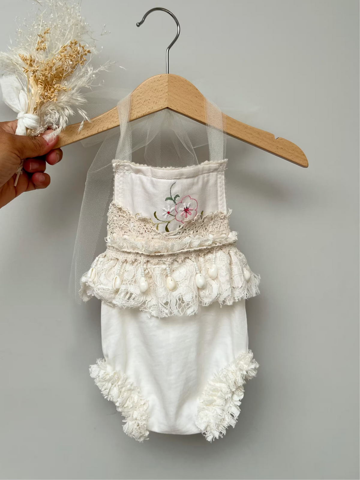 A cream baby romper with delicate lace trim, a small floral embroidery on the chest, and ruffled details, hanging on a wooden hanger with a sheer veil. A hand holding dried flowers is visible on the left against a plain gray background.