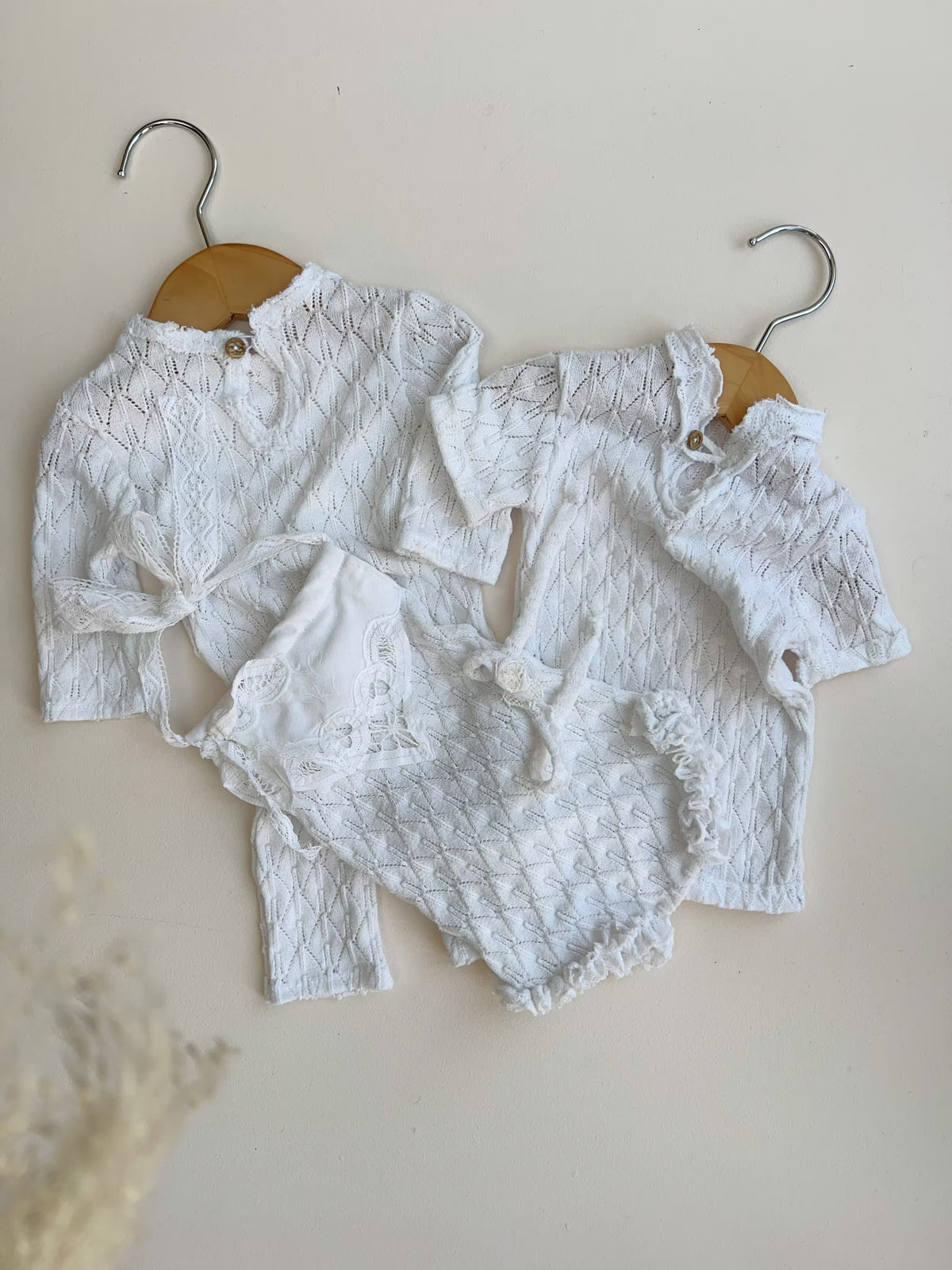 A three-piece white baby clothing set with textured lace and ruffle details, displayed on wooden hangers against a warm beige background.