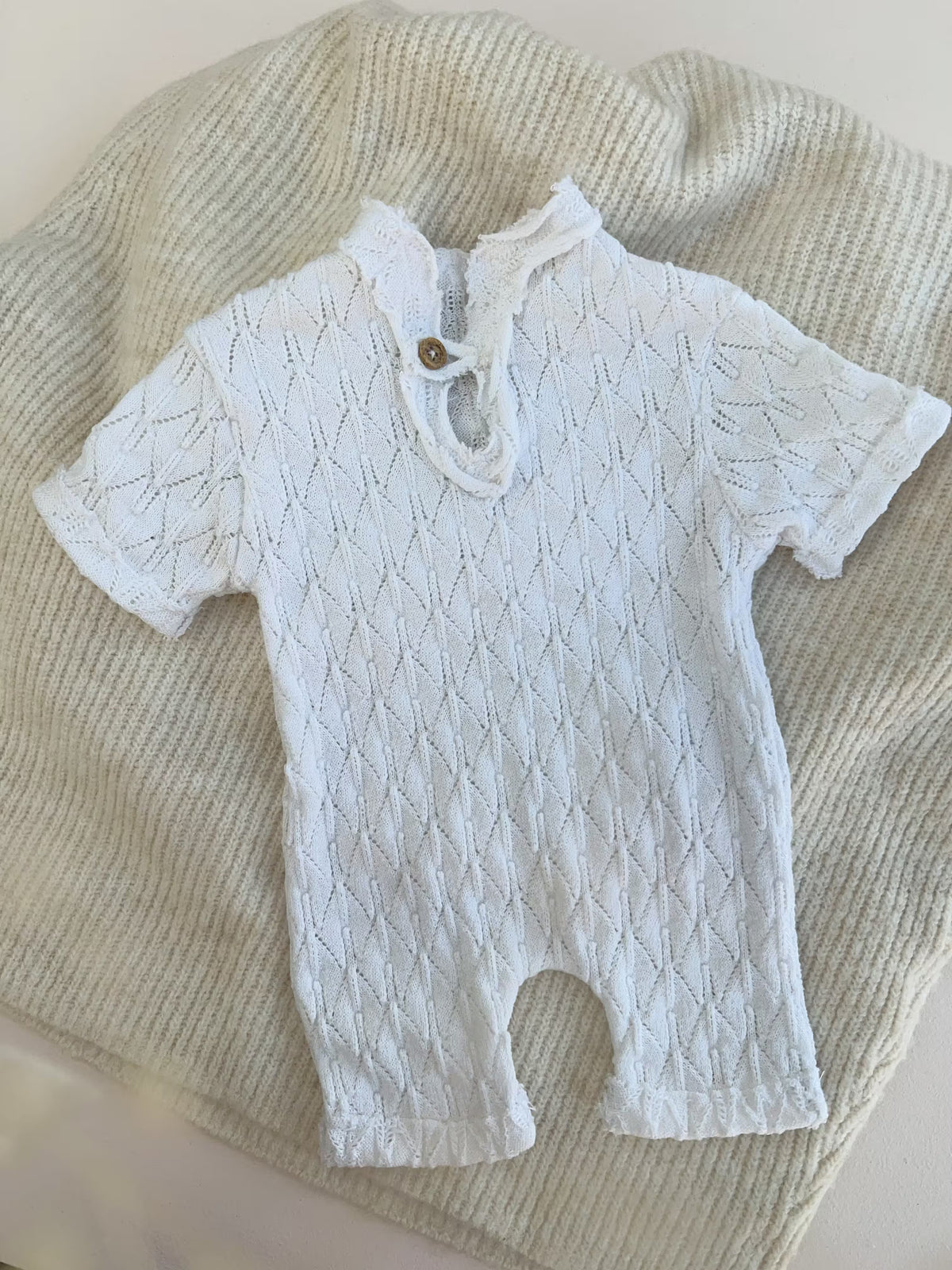 A white short-sleeve baby romper with a textured knit pattern and a small wooden button closure at the collar, laid flat on a cream-colored ribbed blanket.