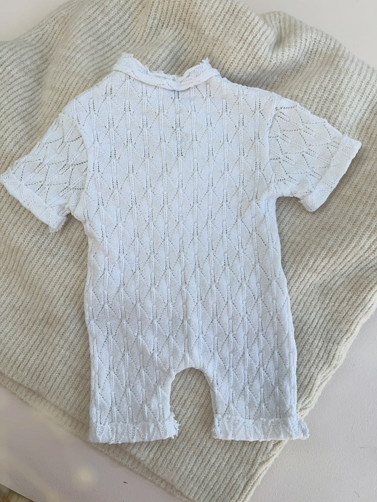 Back of a  white short-sleeve baby romper with a textured diamond knit pattern, laid flat on a cream-colored ribbed blanket.