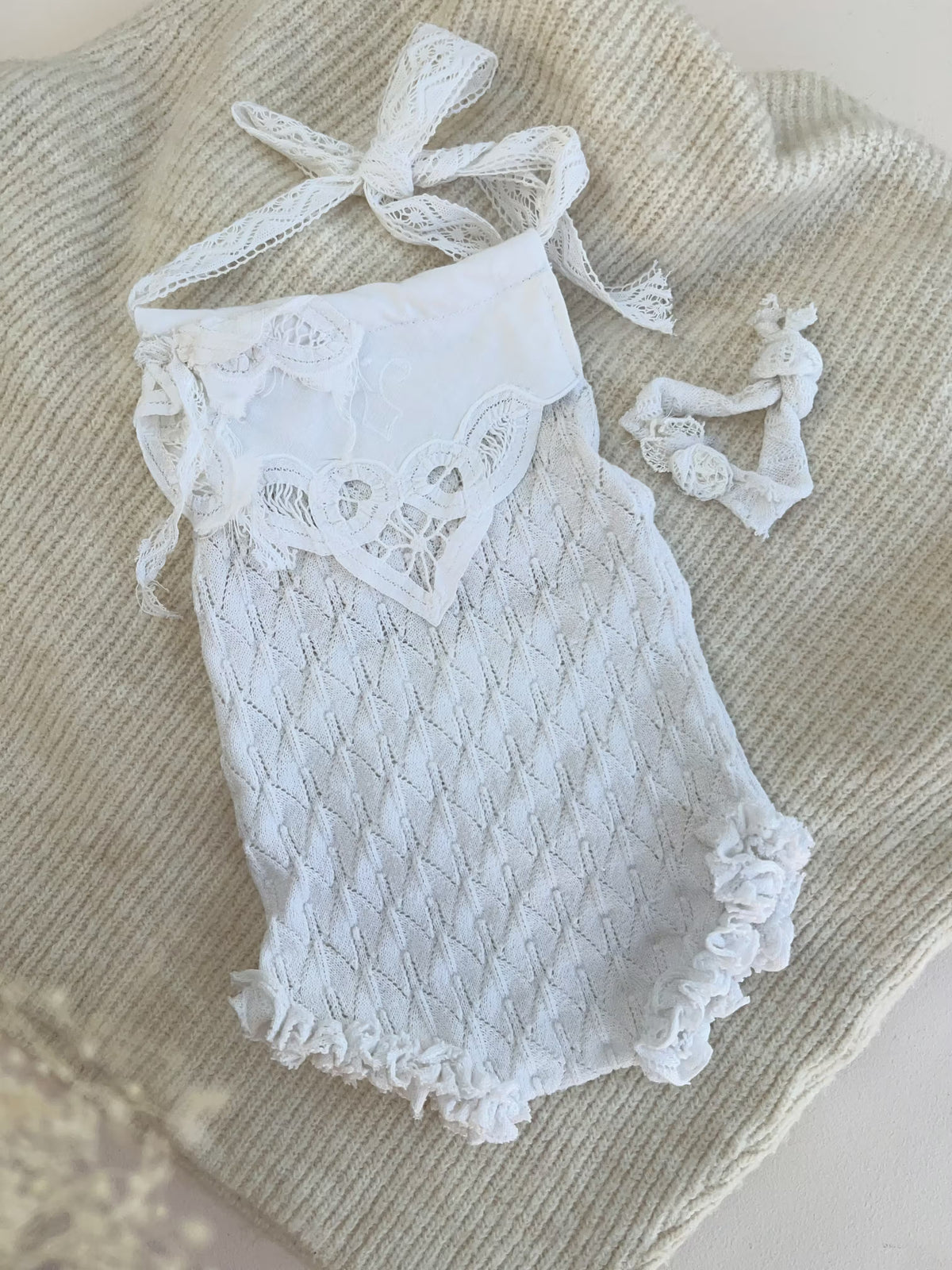White lace baby bodysuit with tie straps and matching headband, laid flat on a beige knit blanket.