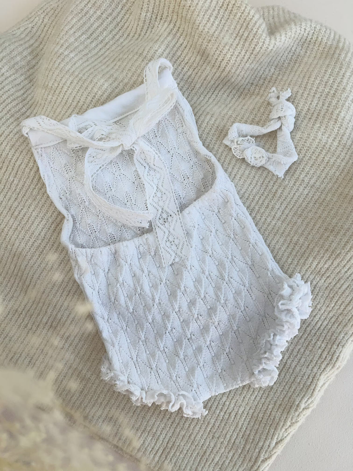 Back detail of the girl romper lace baby bodysuit with ruffled trim, laid flat on a beige knit blanket with a matching white headband nearby.