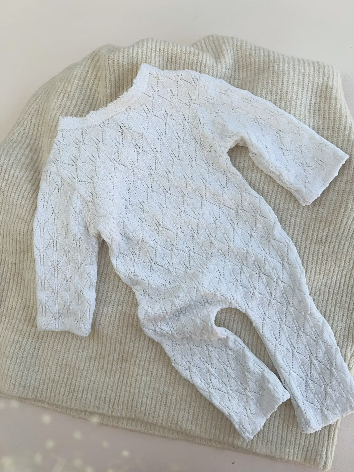 back of a  white long-sleeve baby romper with a textured knit pattern, laid flat on top of a cream ribbed blanket.