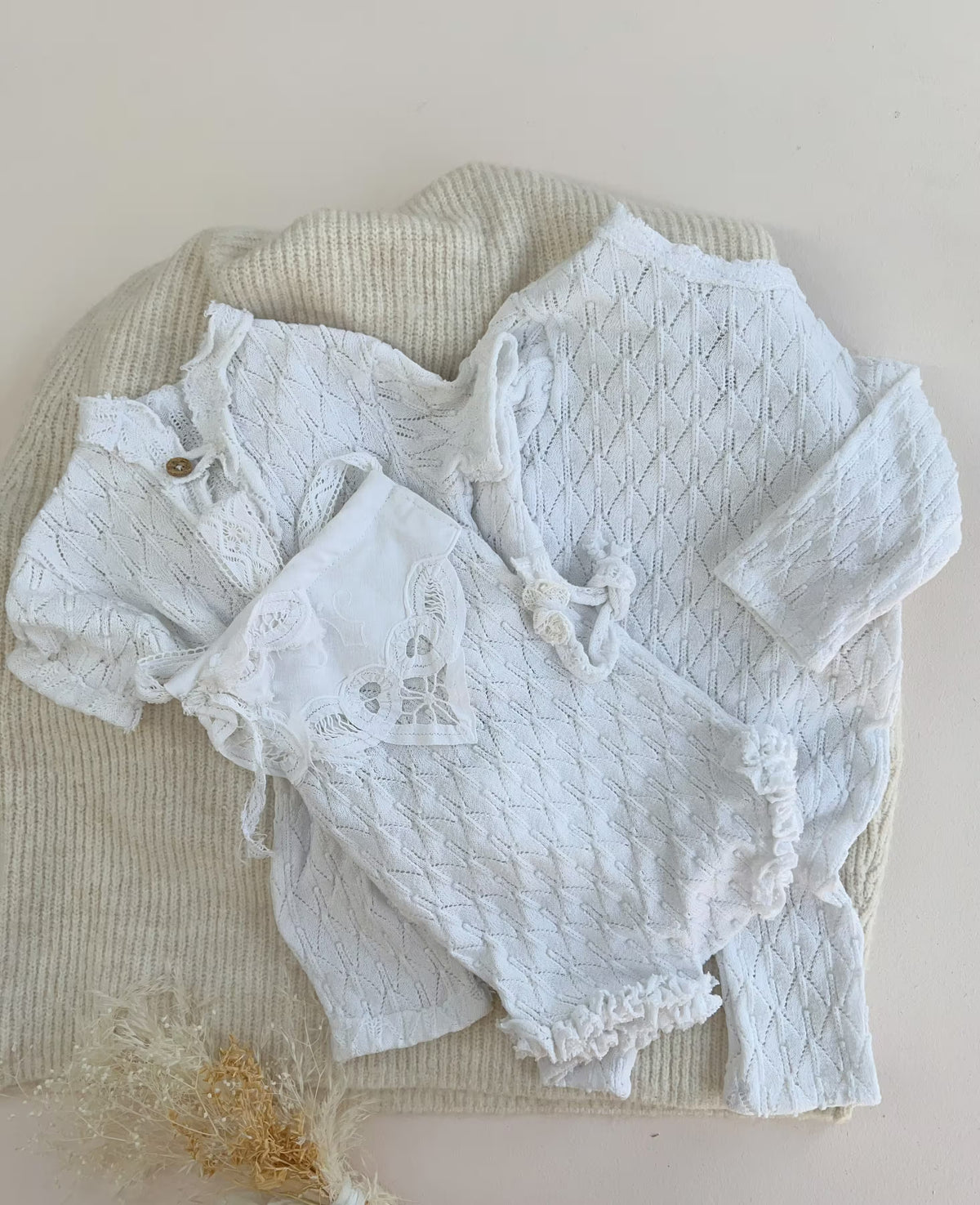A set of white baby clothing, including a long-sleeve textured romper and matching lace-trimmed bodysuit, styled on top of a cream ribbed knit blanket.